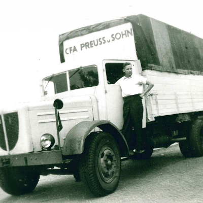 Alter LKW mit der Aufschrift VFA Preuss u. Sohn über dem Fahrerhaus. Auf der Außenseite des Fahrerhauses steht ein Mann und lenkt den LKW von draußen.