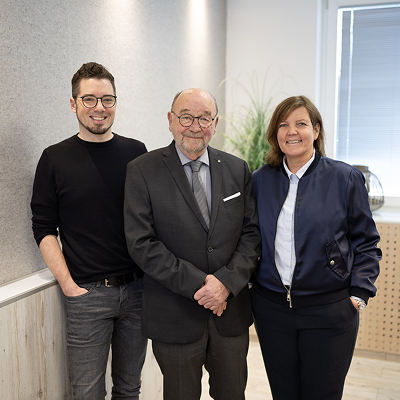 3 Personen der Preuss Familie stehen nebeneinander. Links steht der Enkel, in der Mitte der Gründer und rechts die Tochter des Gründers. Die Personen stehen dicht beieinander